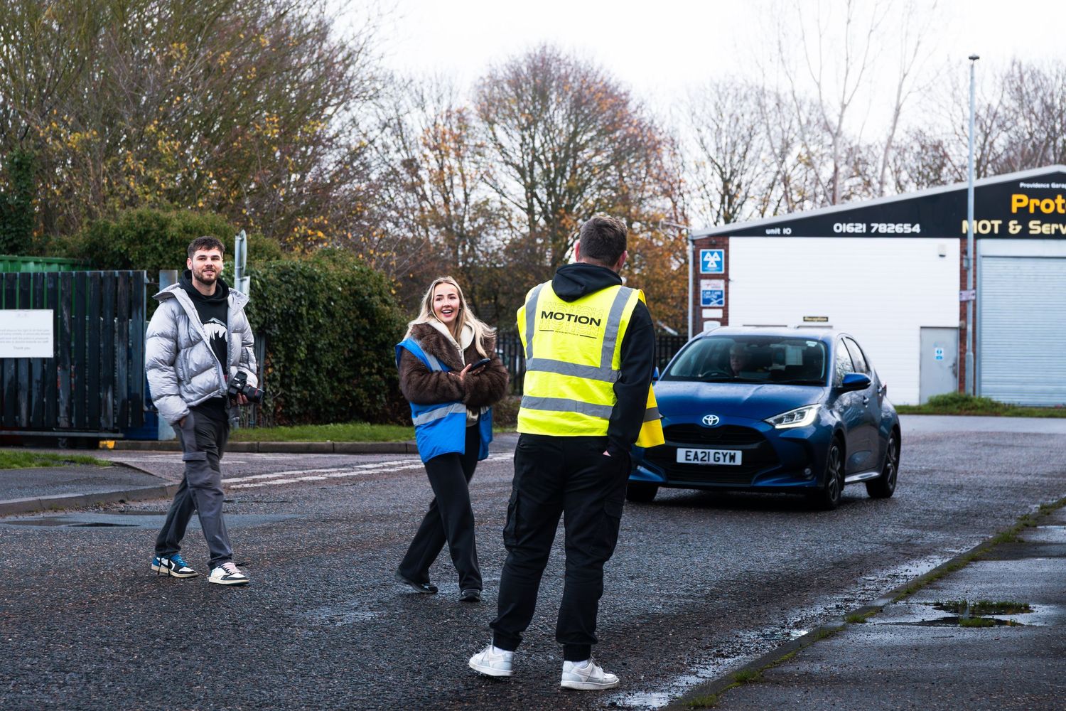 A marshal in a hi-vis vest greets two attendees at an outdoor automotive meet event.