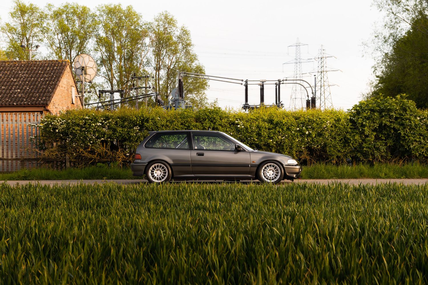 Modified grey Honda Civic hatchback with BBS wheels on a country lane beside green hedges.