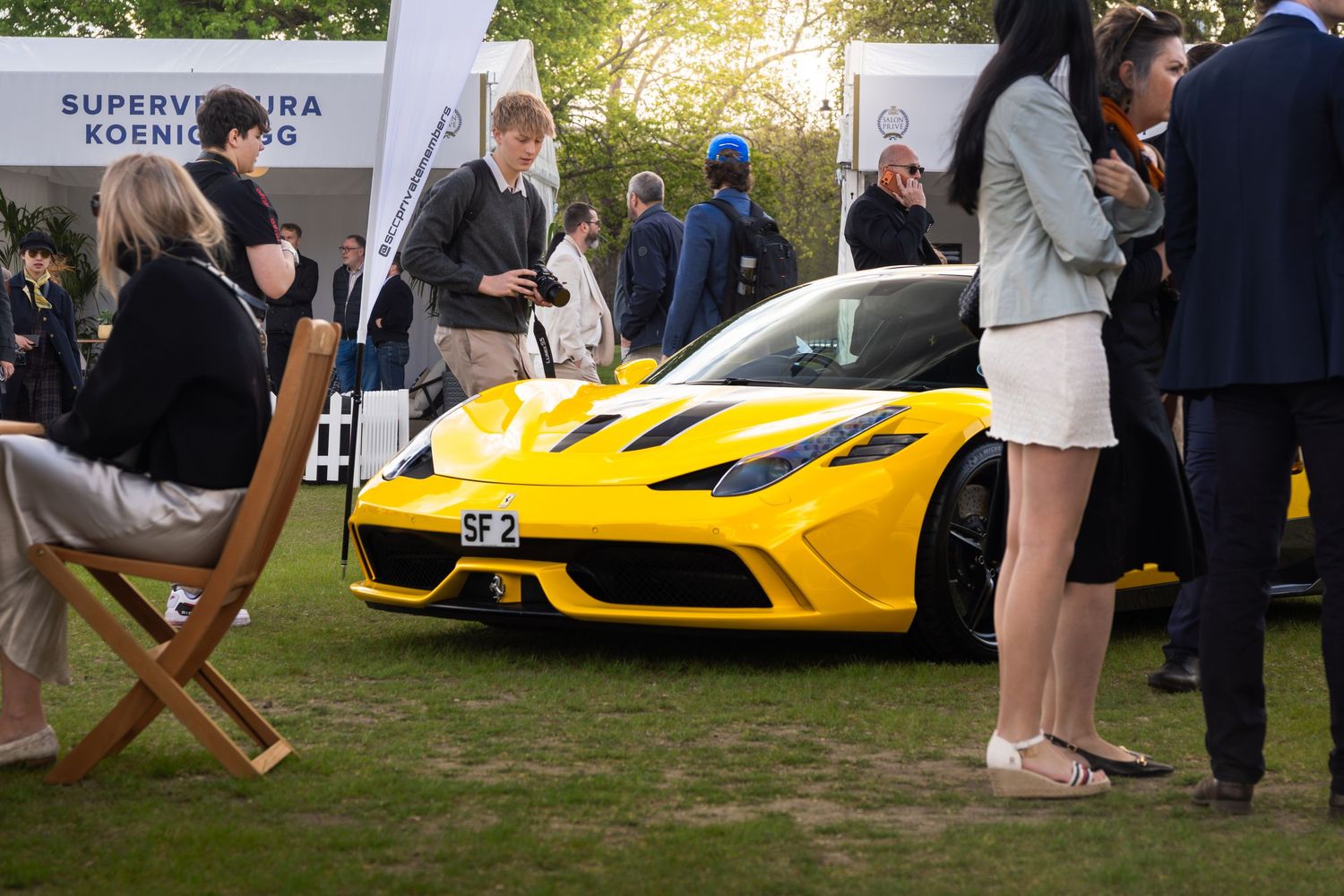 Yellow Ferrari SF90 on display at an outdoor luxury car show with elegantly dressed attendees.