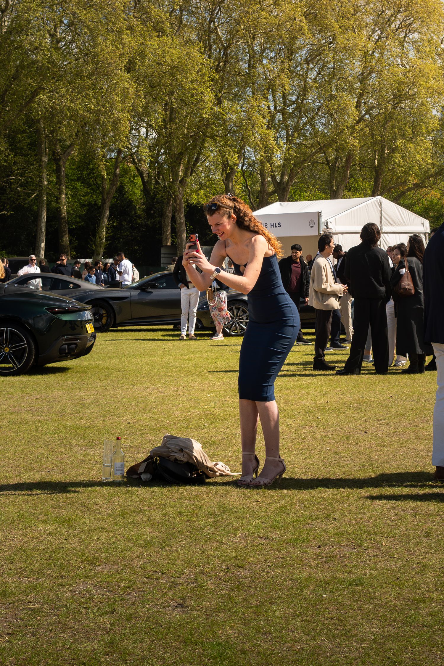 Woman in blue dress photographing cars at an outdoor luxury car show on a sunny day.