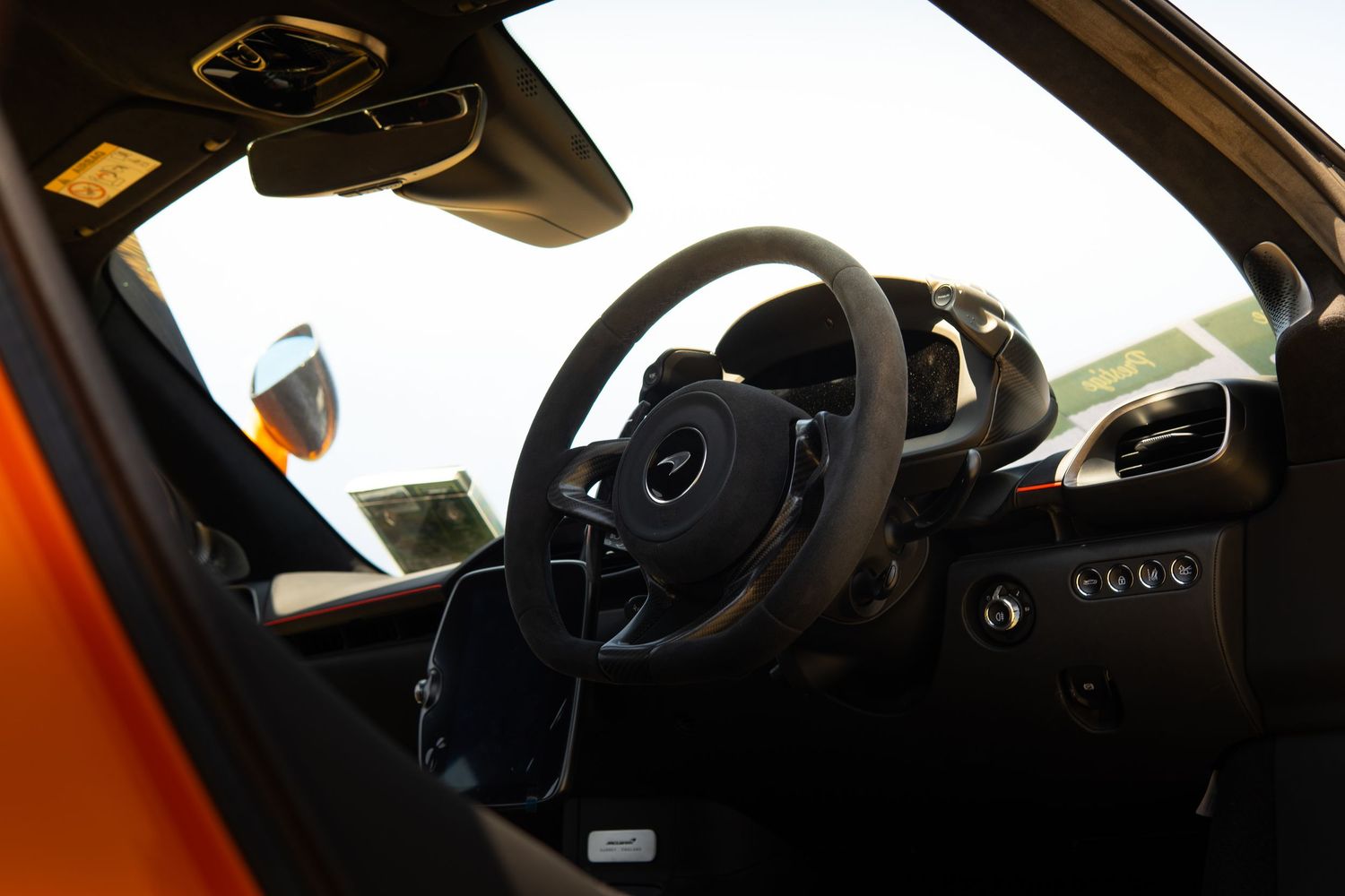 Interior view of an orange McLaren supercar showing steering wheel and carbon fiber dashboard.