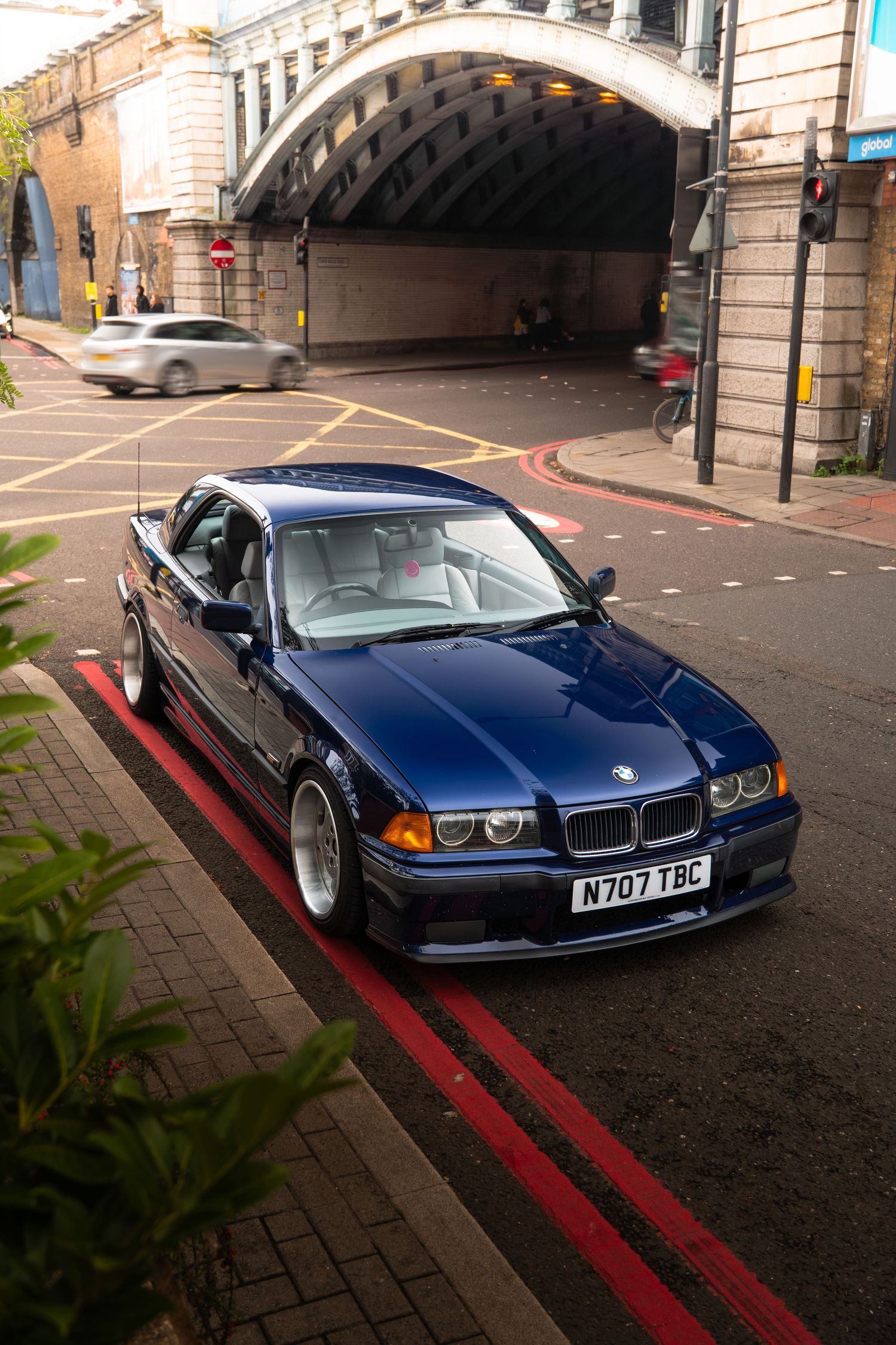 A lowered blue BMW E36 coupe on silver wheels parked on red lines beneath a brick arch bridge.