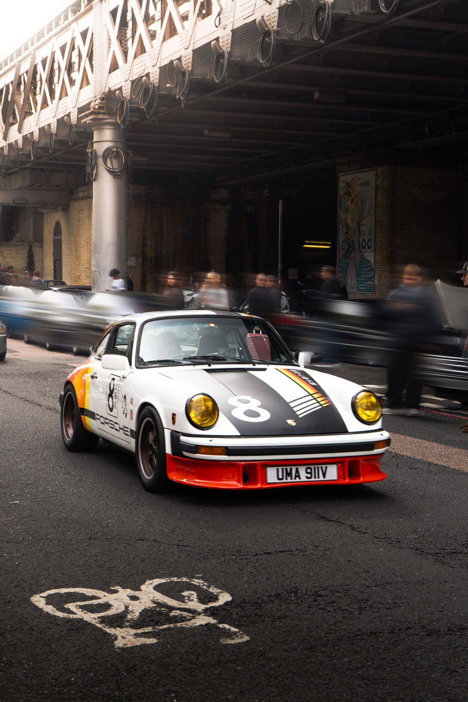 A classic white Porsche 911 RSR with racing livery and yellow headlights driving under a bridge.
