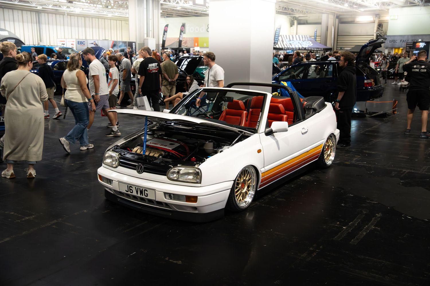 White VW Golf convertible with red interior, gold wheels, and open hood at an indoor car show.