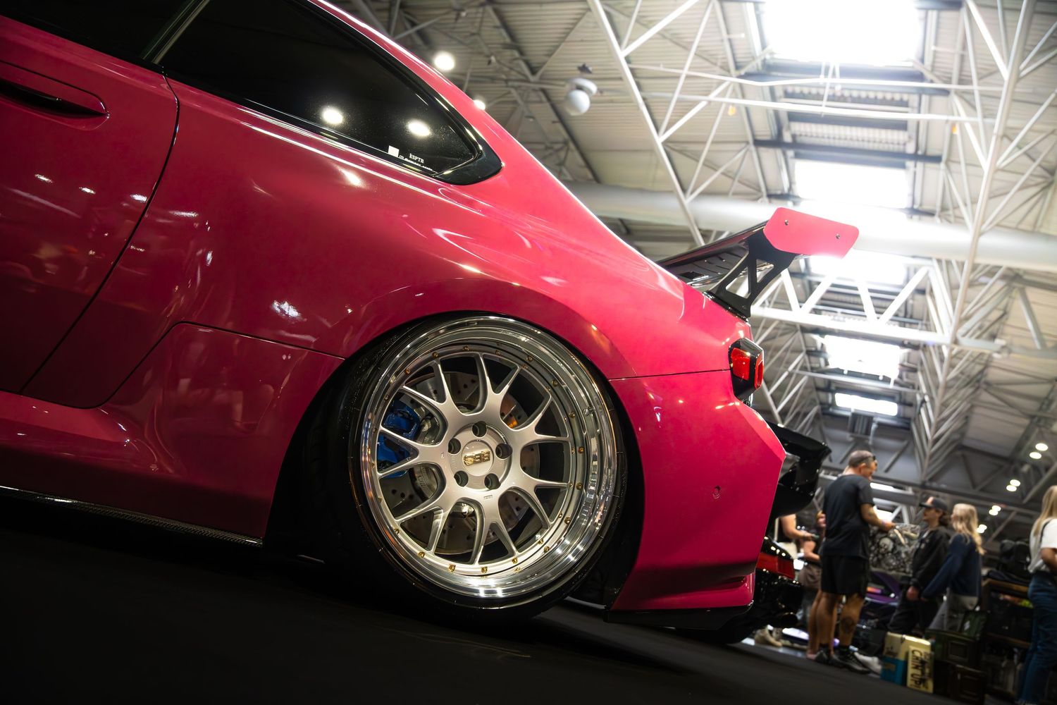 Lowered pink modified car with large rear spoiler and silver alloy wheels at an indoor car show.