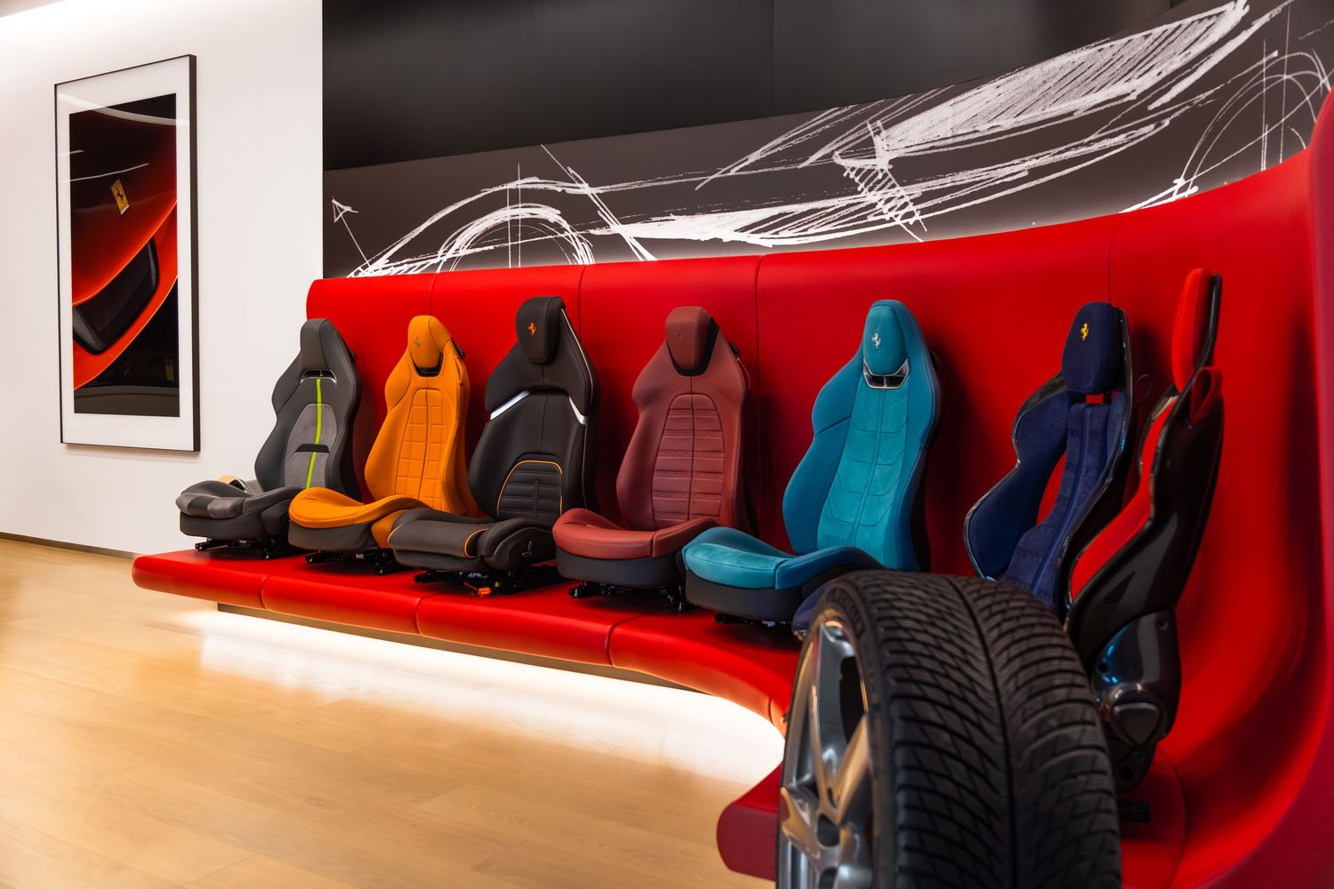 Colorful Ferrari car seats display in red showroom with various upholstery options and tires on wooden floor.