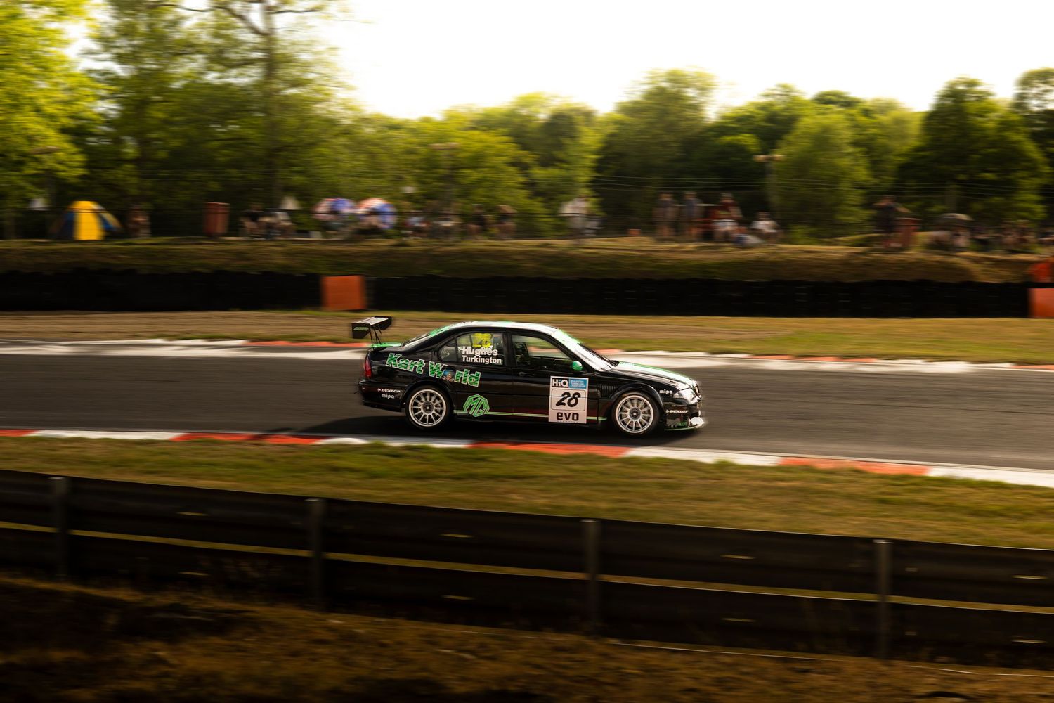 Black racing sedan number 44 cornering at speed on a circuit track lined with trees and spectators.