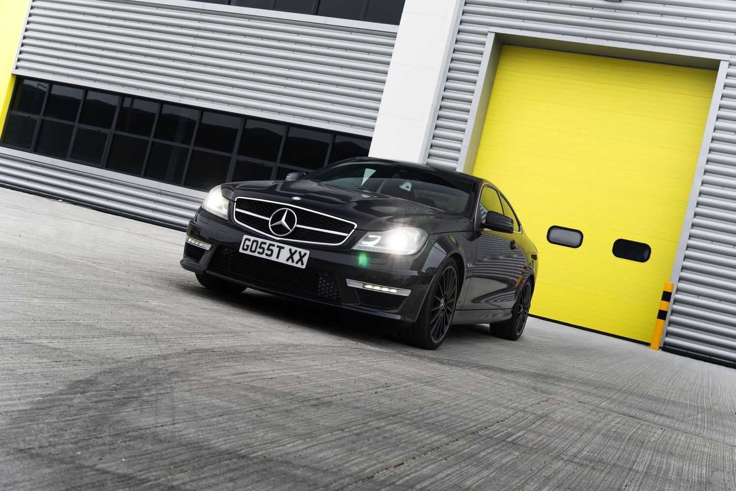 Black Mercedes-AMG C63 coupe driving past bright yellow garage doors at an industrial facility.