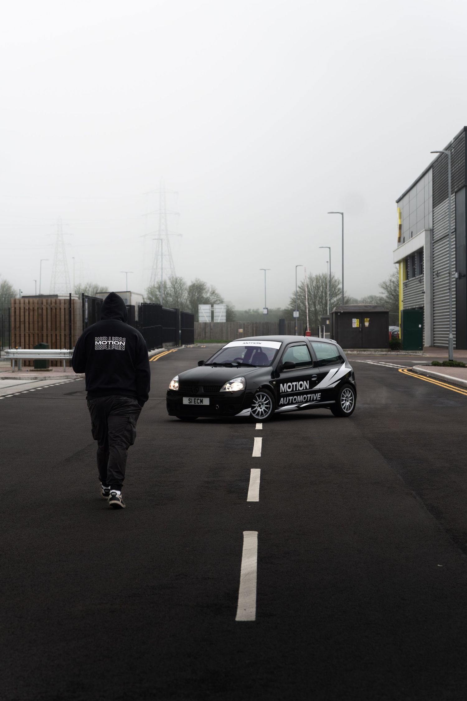 Motion Automotive branded person walking along a misty road toward a parked black Renault Clio on an industrial estate.