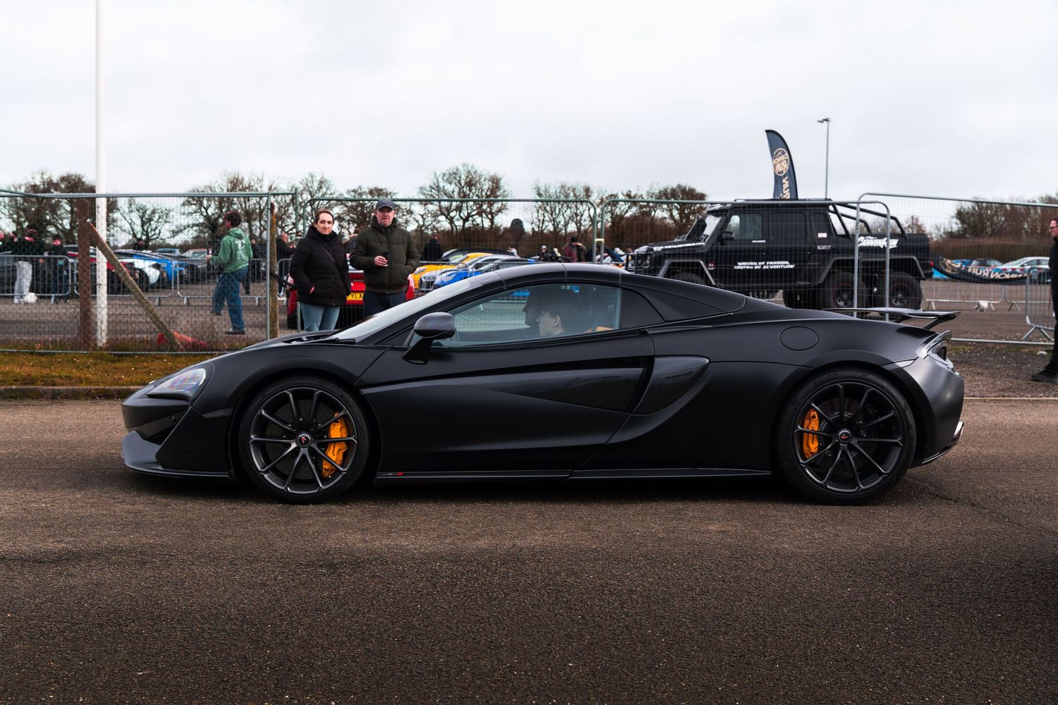 A matte black McLaren 570S supercar parked at an outdoor car meet event on an overcast day.