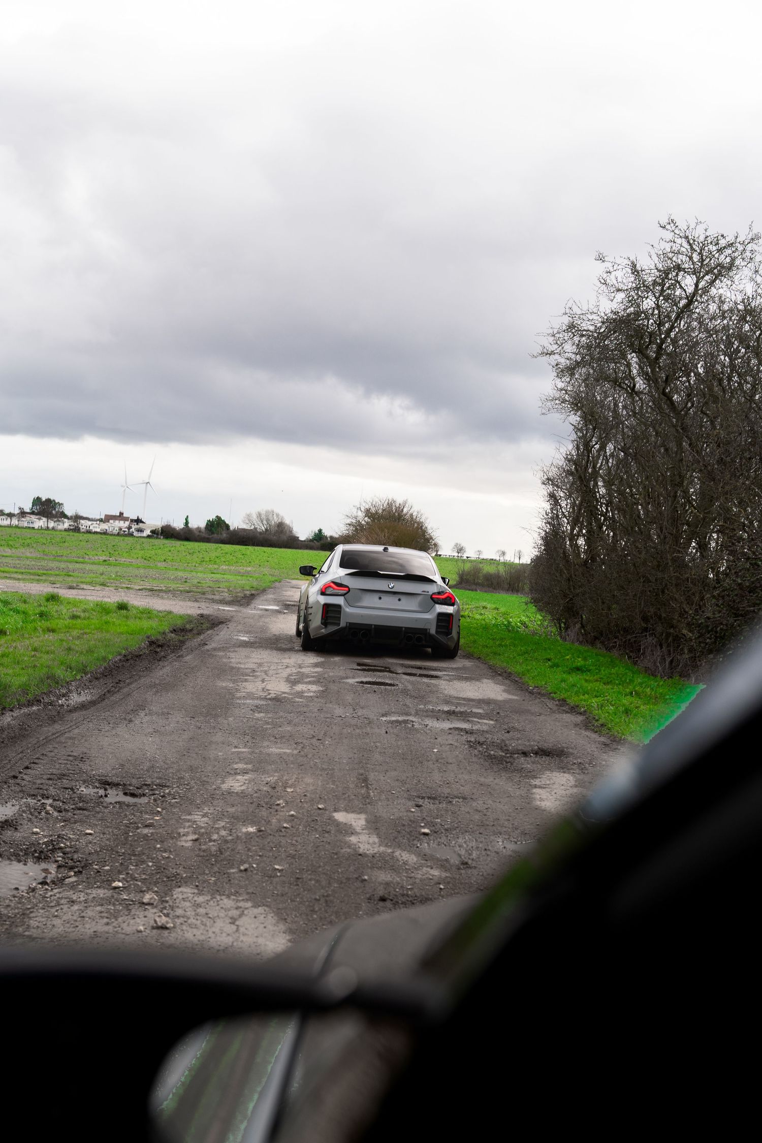 Silver BMW M3 driving away on a rough rural road flanked by green fields and bare trees.