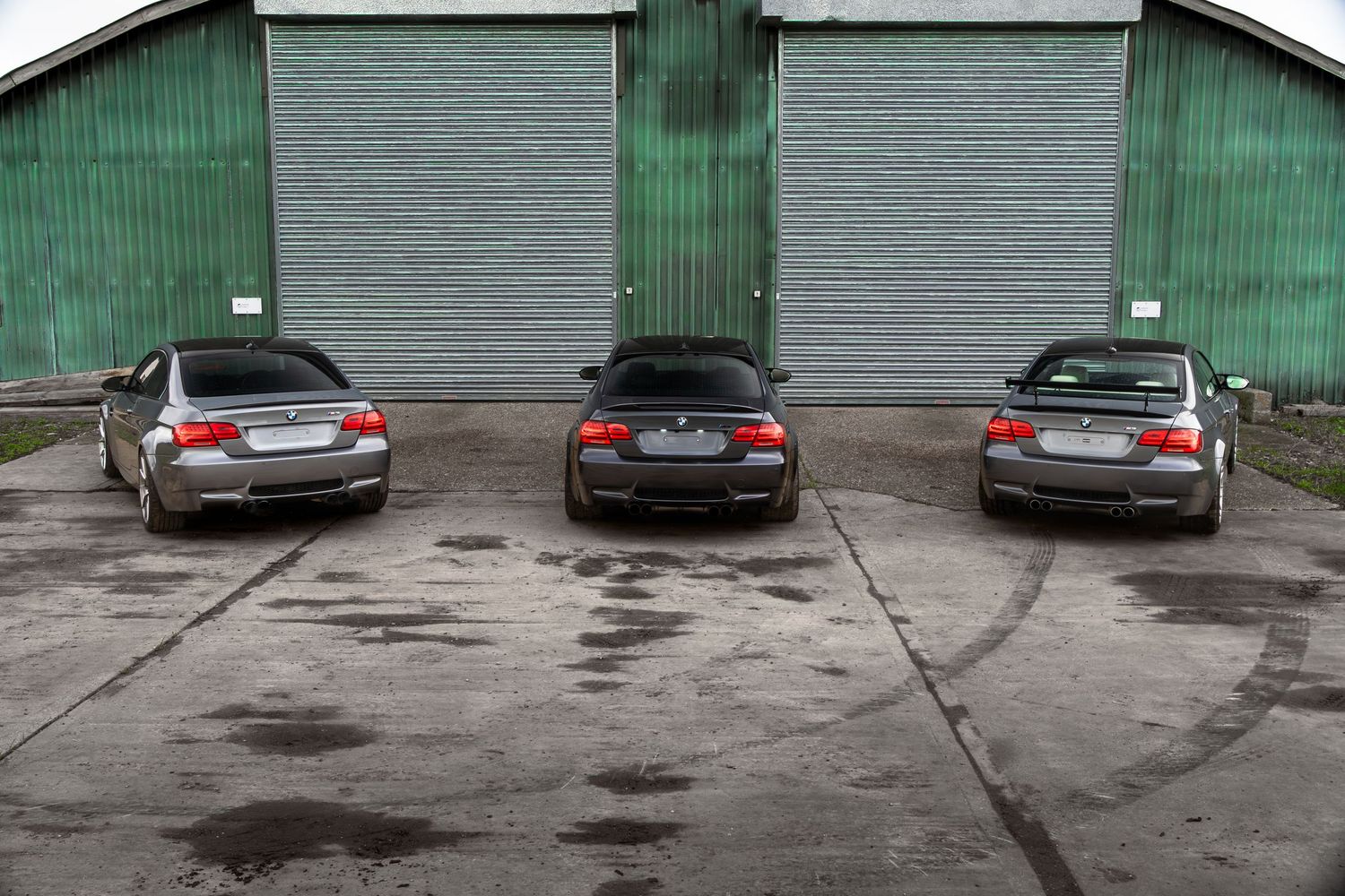 Rear view of three BMW M3 coupes in dark grey finish parked in front of green roller doors.