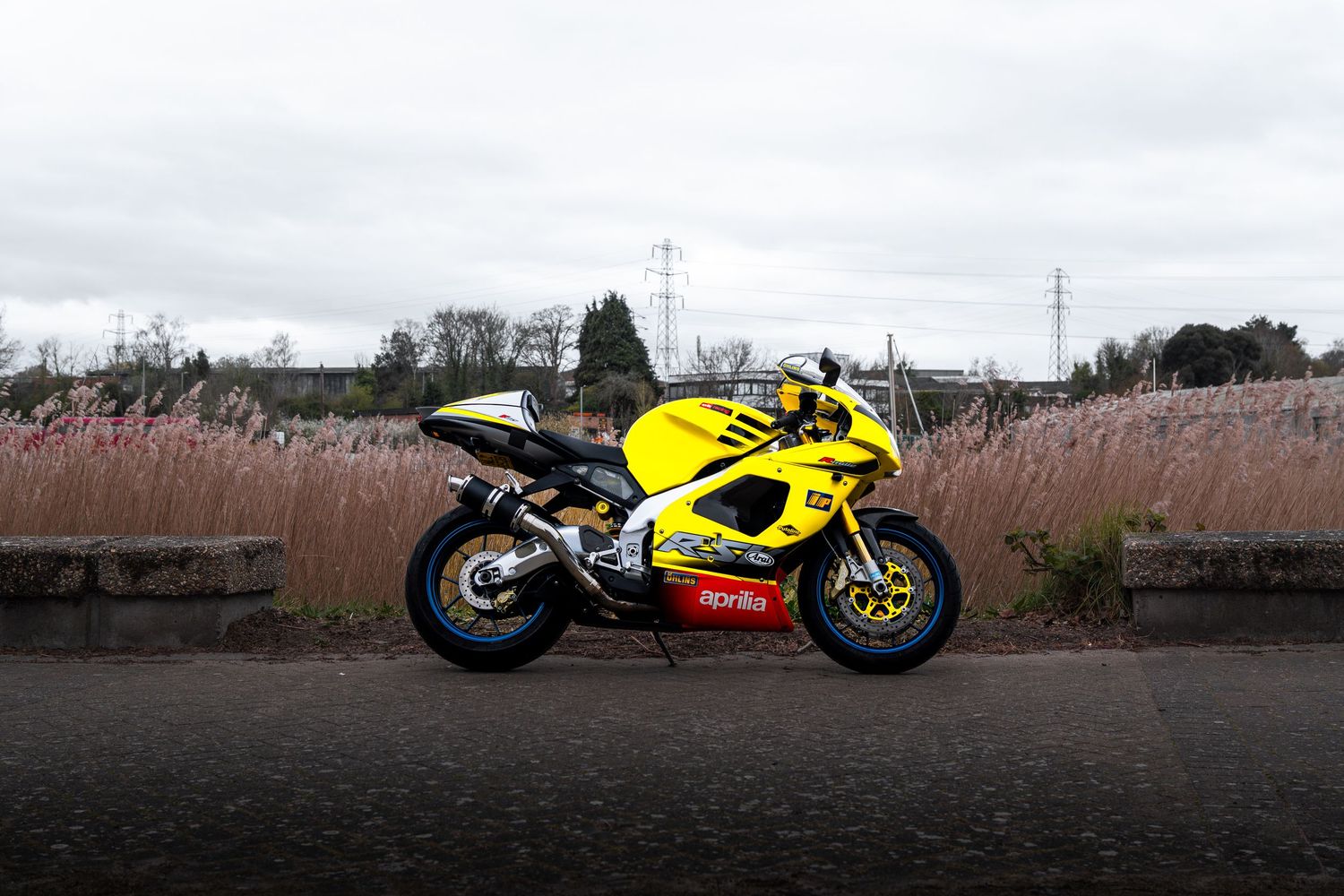 Yellow Aprilia RSV superbike parked beside tall grass with overcast sky in background.