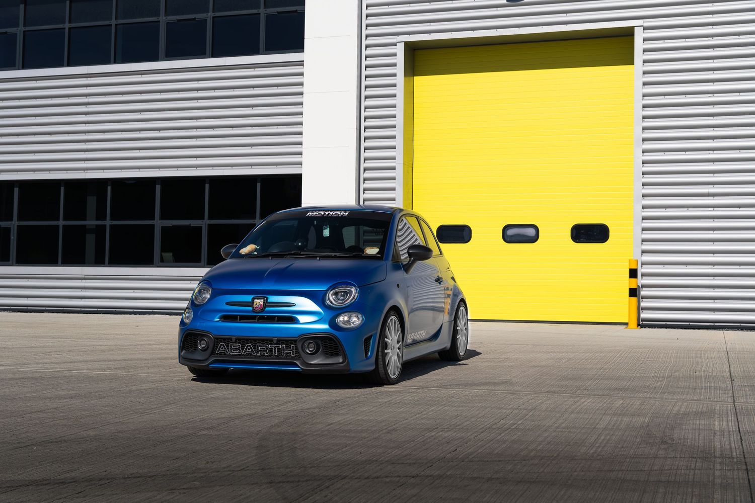 Blue Abarth 500 sports car posed in front of bright yellow roller shutter garage doors.