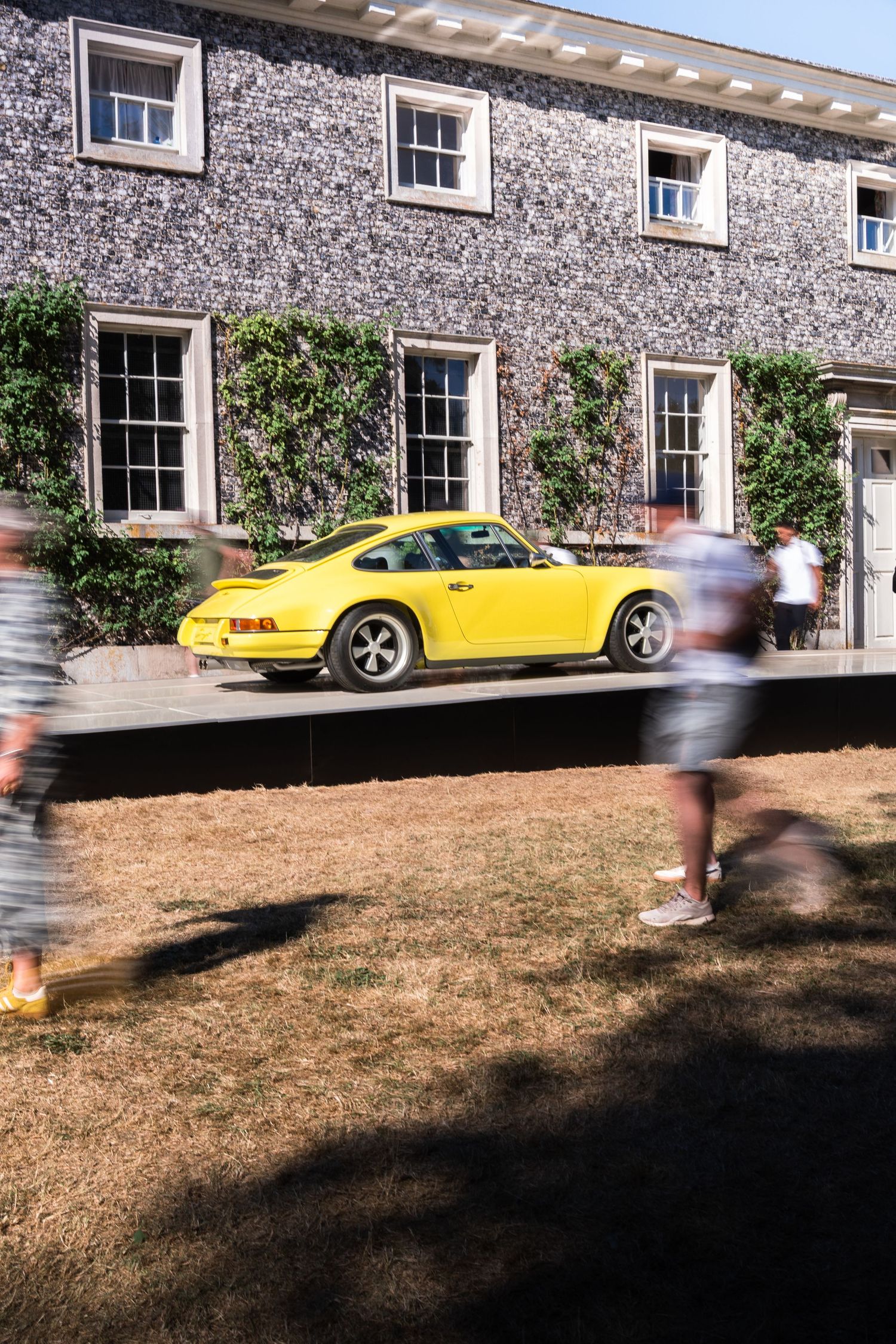 Bright yellow Porsche 911 parked outside a stone building with ivy, as blurred pedestrians walk past on a sunny day.