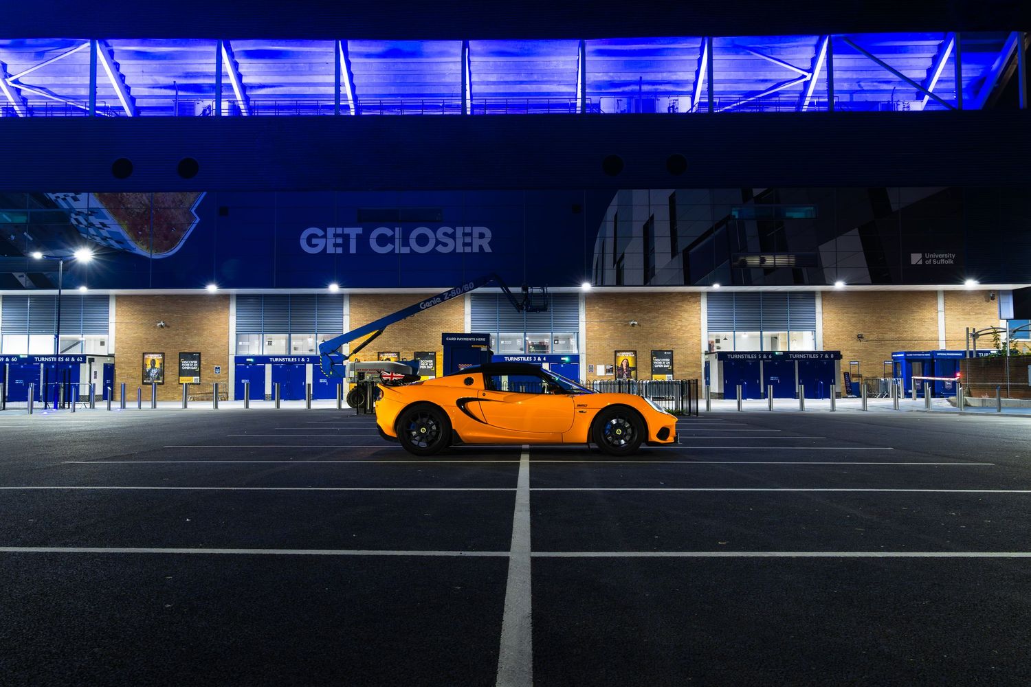 Wide shot of orange Lotus Elise in night-time car park beside blue-lit stadium facade with GET CLOSER sign.