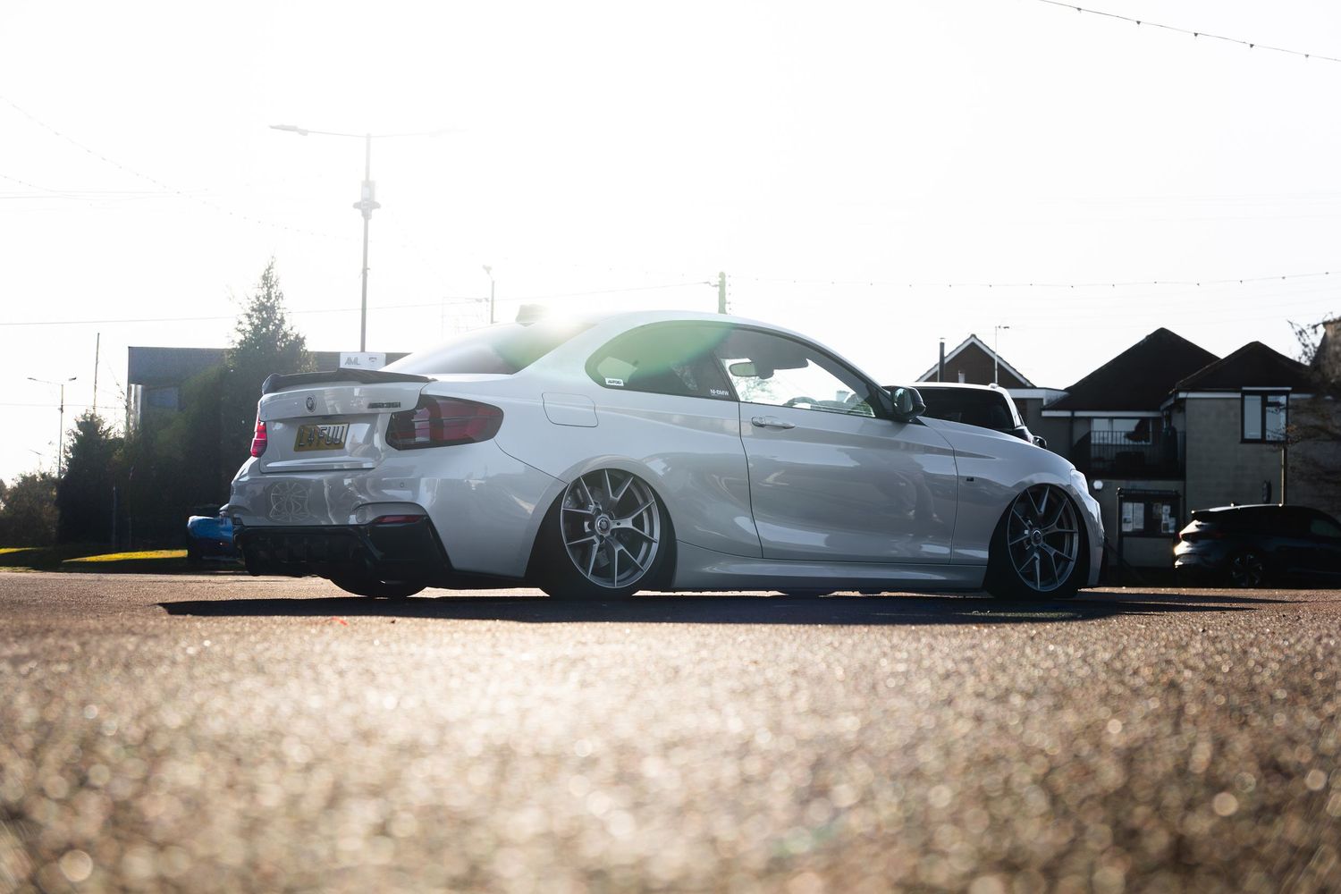 Lowered white BMW 2 Series coupe with aftermarket wheels parked in golden sunlight.