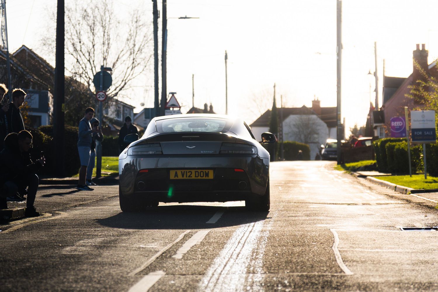 Aston Martin sports car driving away on a sunny UK village road.