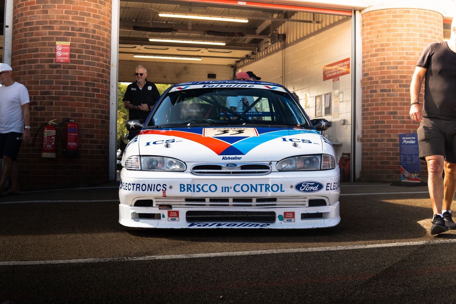 White Ford Mondeo racing car with Brisco branding parked outside pit garage at circuit.