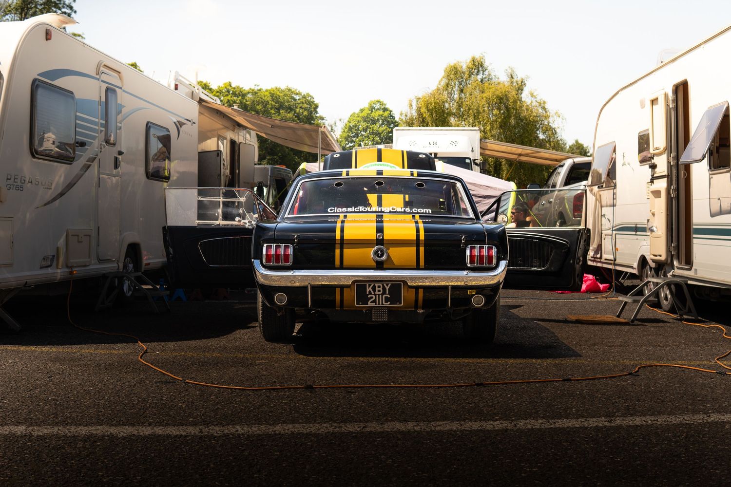 Classic Ford Mustang with yellow racing stripes parked between caravans in paddock area.