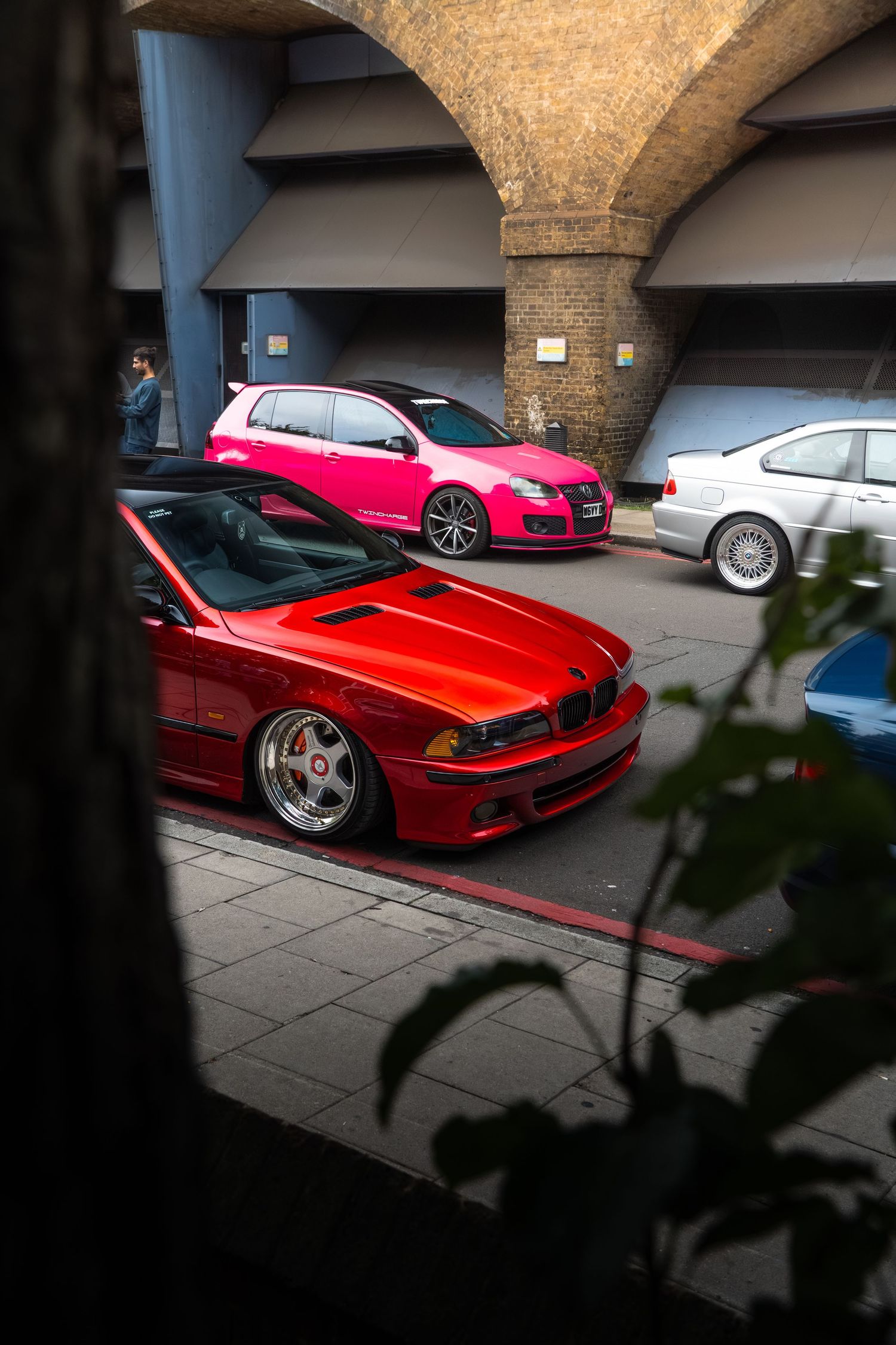 Red BMW E39 and pink VW Golf parked under stone arches at a car meet.