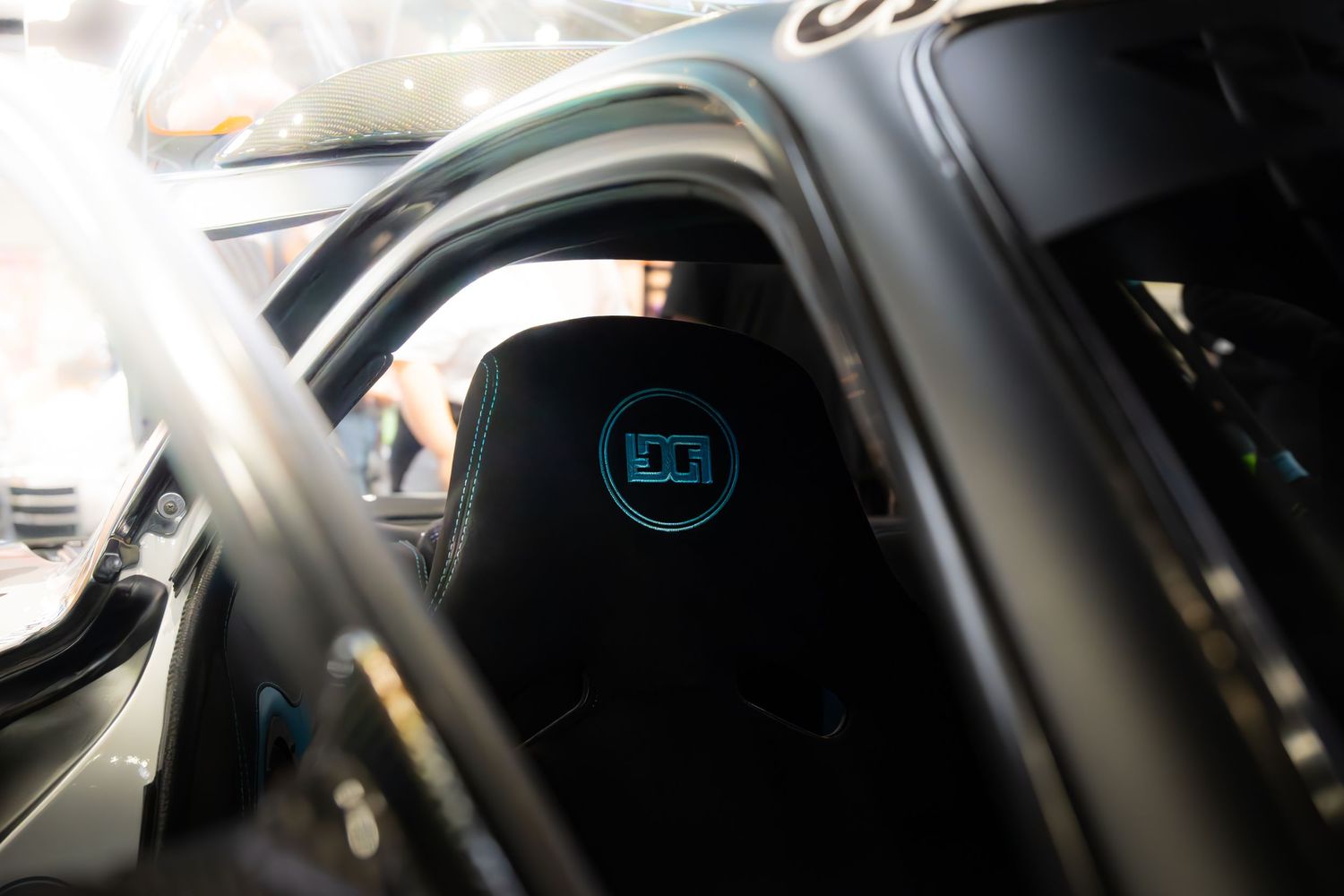 Interior view of a modified car showing black racing seat with blue logo emblem through roll cage.