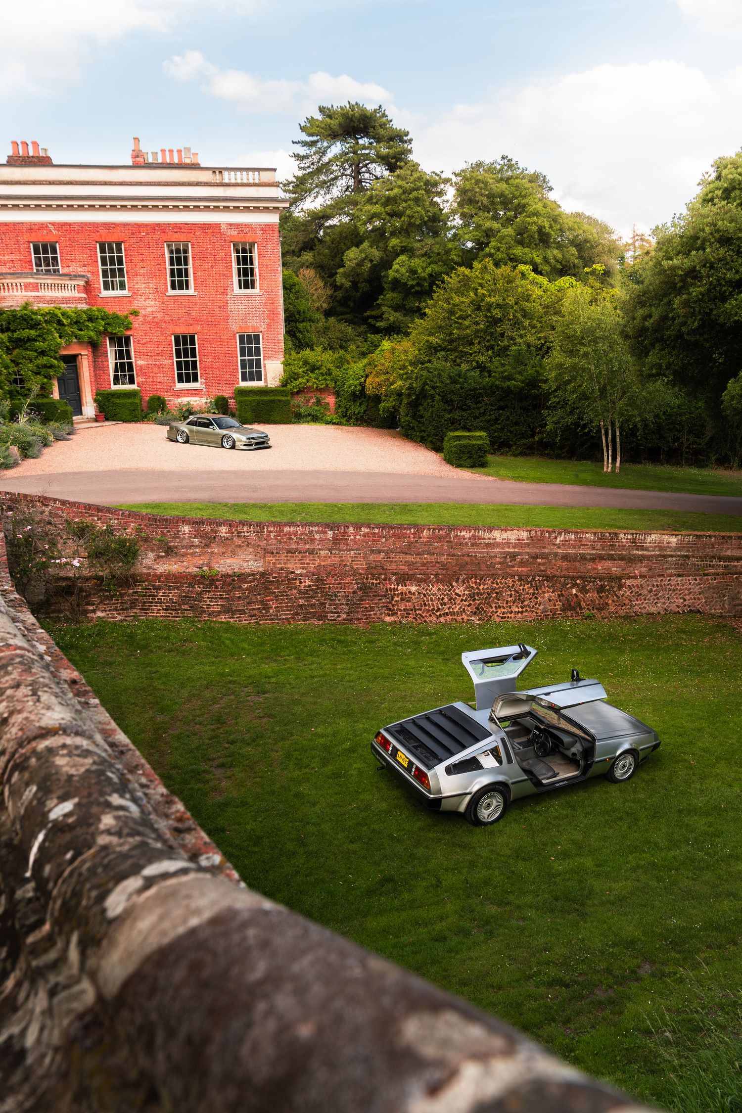 A DeLorean DMC-12 with gull-wing doors open on green lawn beside a red brick Georgian country house.