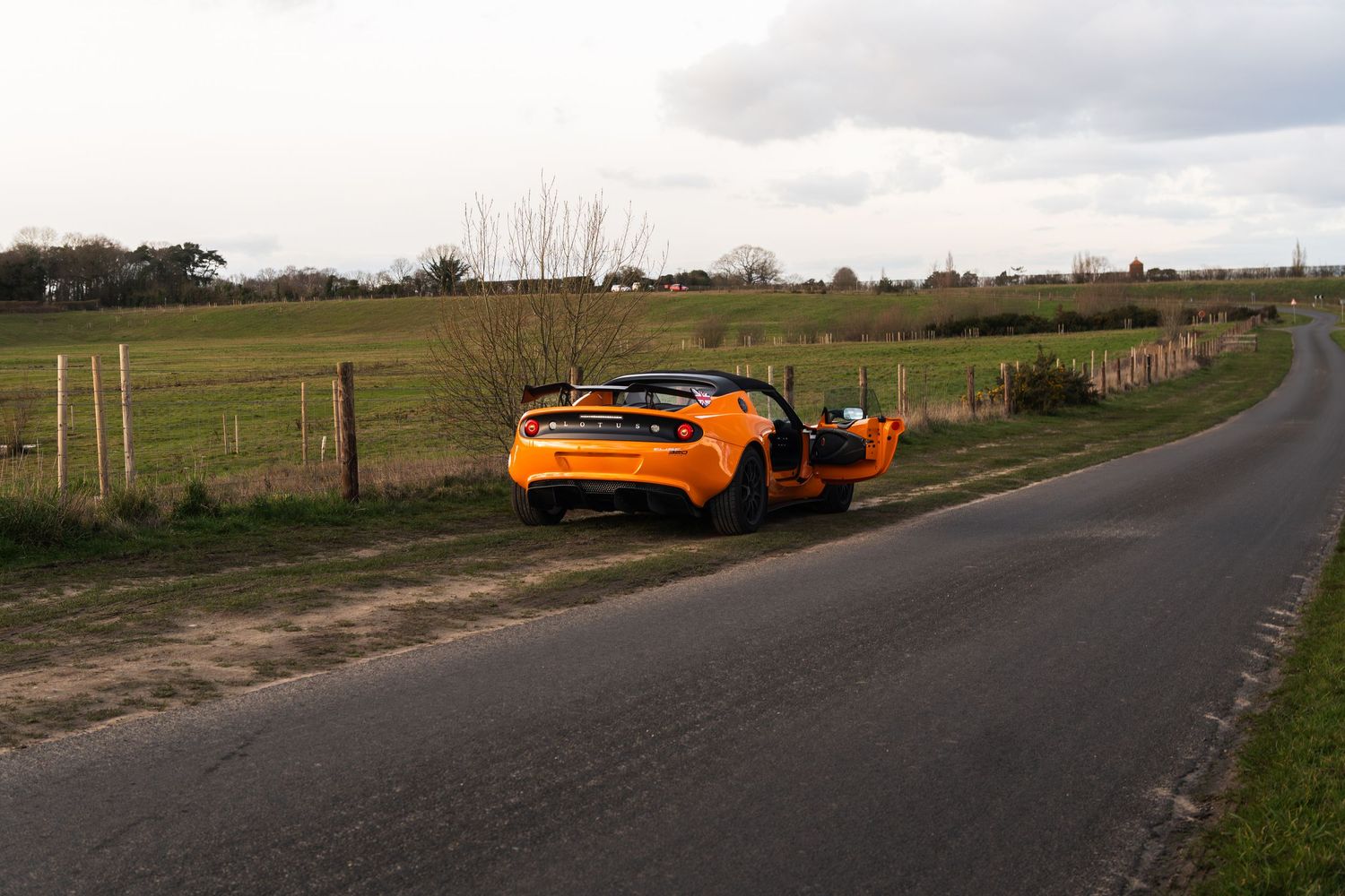 Bright orange Lotus Elise with doors open parked on winding rural countryside road.