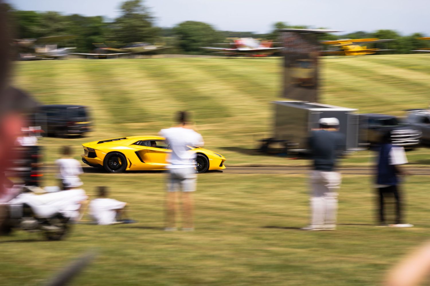 Yellow Lamborghini Aventador speeding across a green field at an outdoor car event with spectators watching.