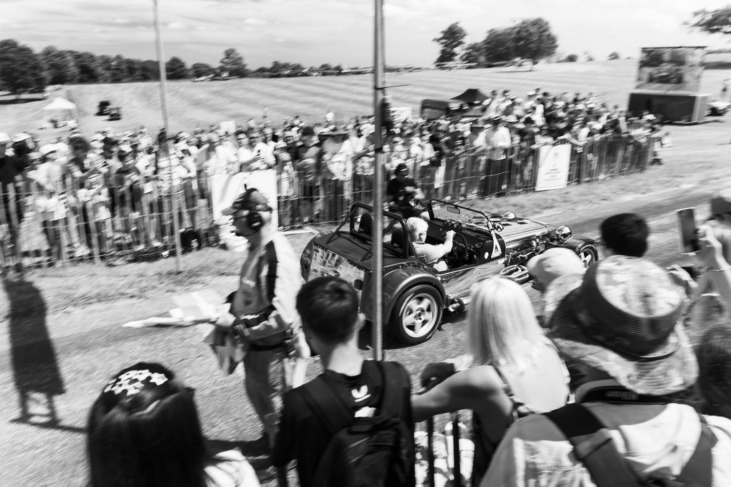 Black and white photo of a vintage race car driving past a large crowd of spectators at an outdoor motorsport event.
