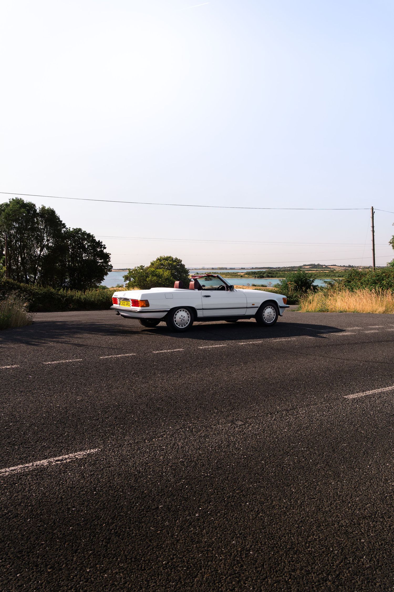 White convertible Mercedes-Benz parked on a coastal road surrounded by greenery and water on a clear day.