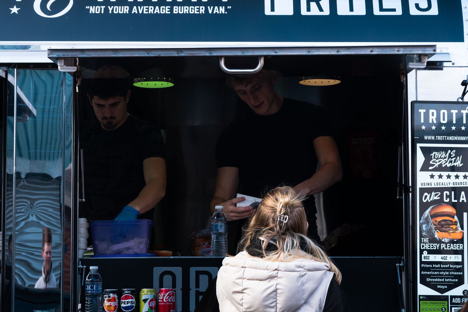 Two vendors serving food from a burger van to a customer at an outdoor automotive event.