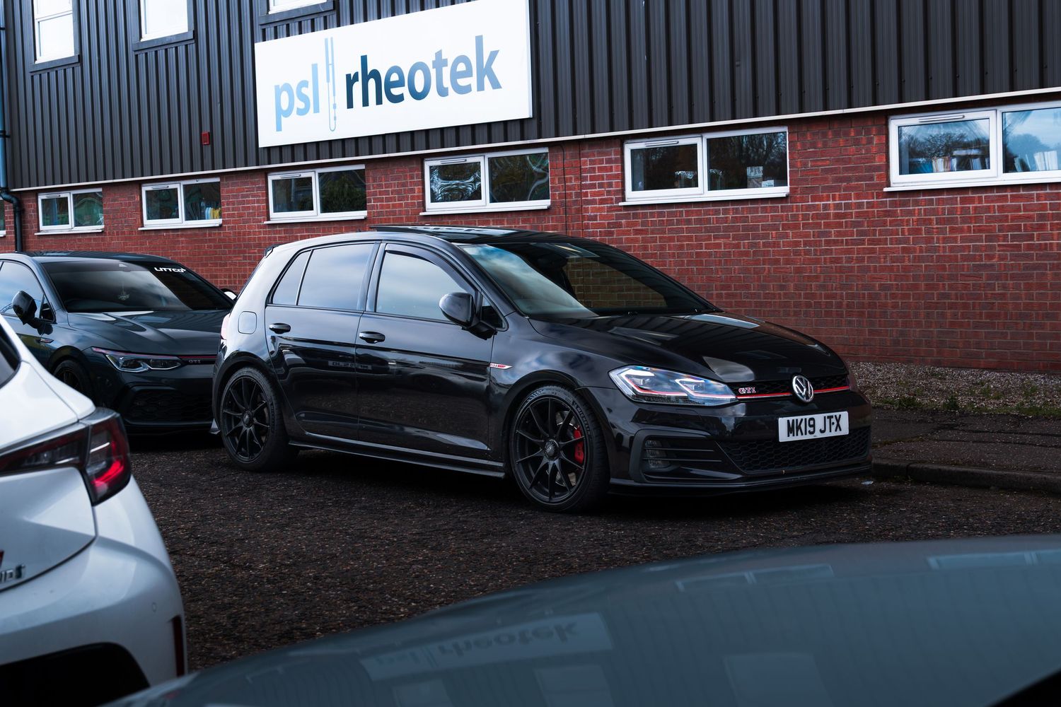Black Volkswagen Golf GTI parked outside PSL Rheotek building on a cloudy day.
