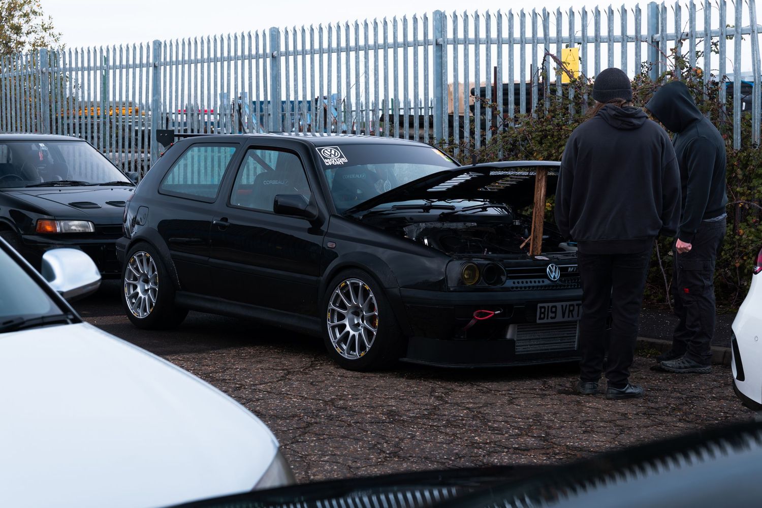 Black VW Golf with hood open being inspected by two men in a car meet parking lot.