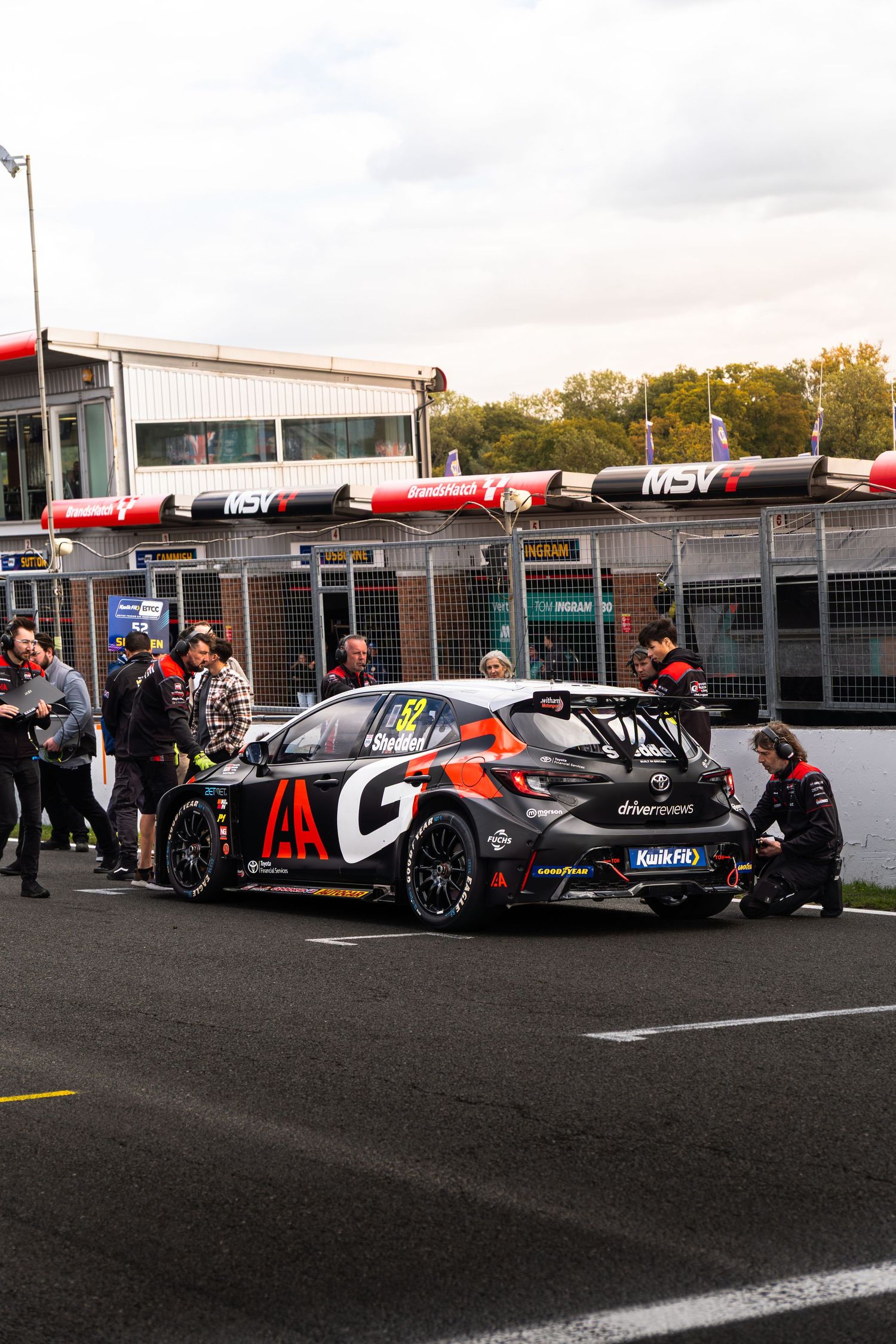 Dark BTCC Toyota GR Corolla race car in pit lane with mechanics working on it at a motorsport circuit.