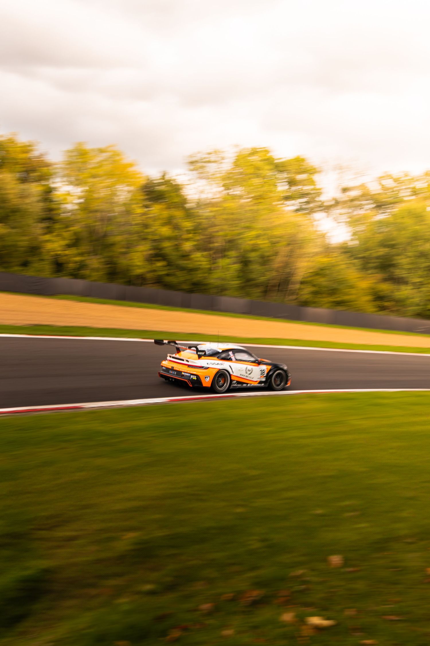 Orange GT race car speeding through a circuit bend with lush green grass and cloudy sky above.