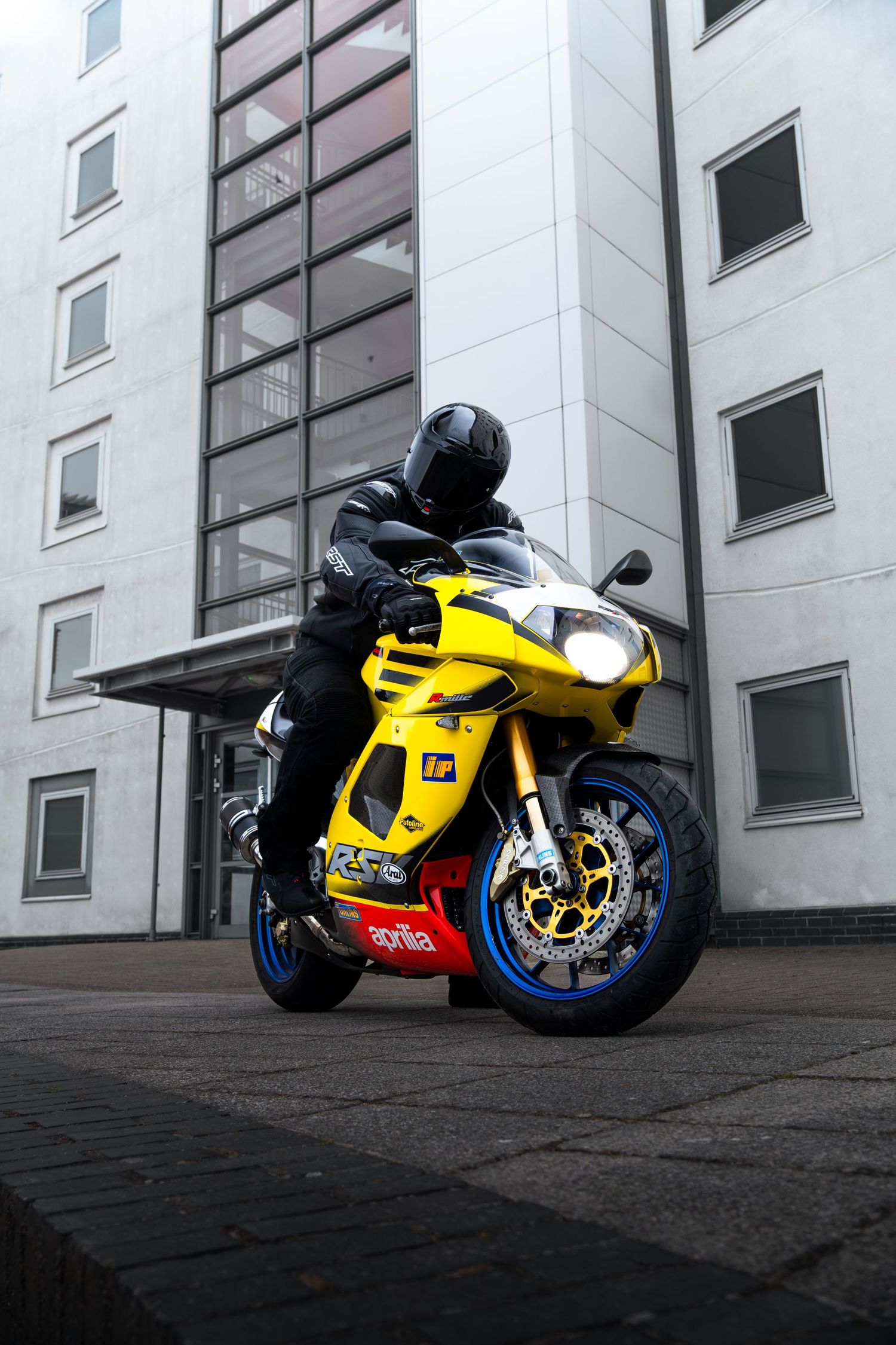 Rider on yellow Aprilia superbike with blue wheels posed outside a modern glass building.