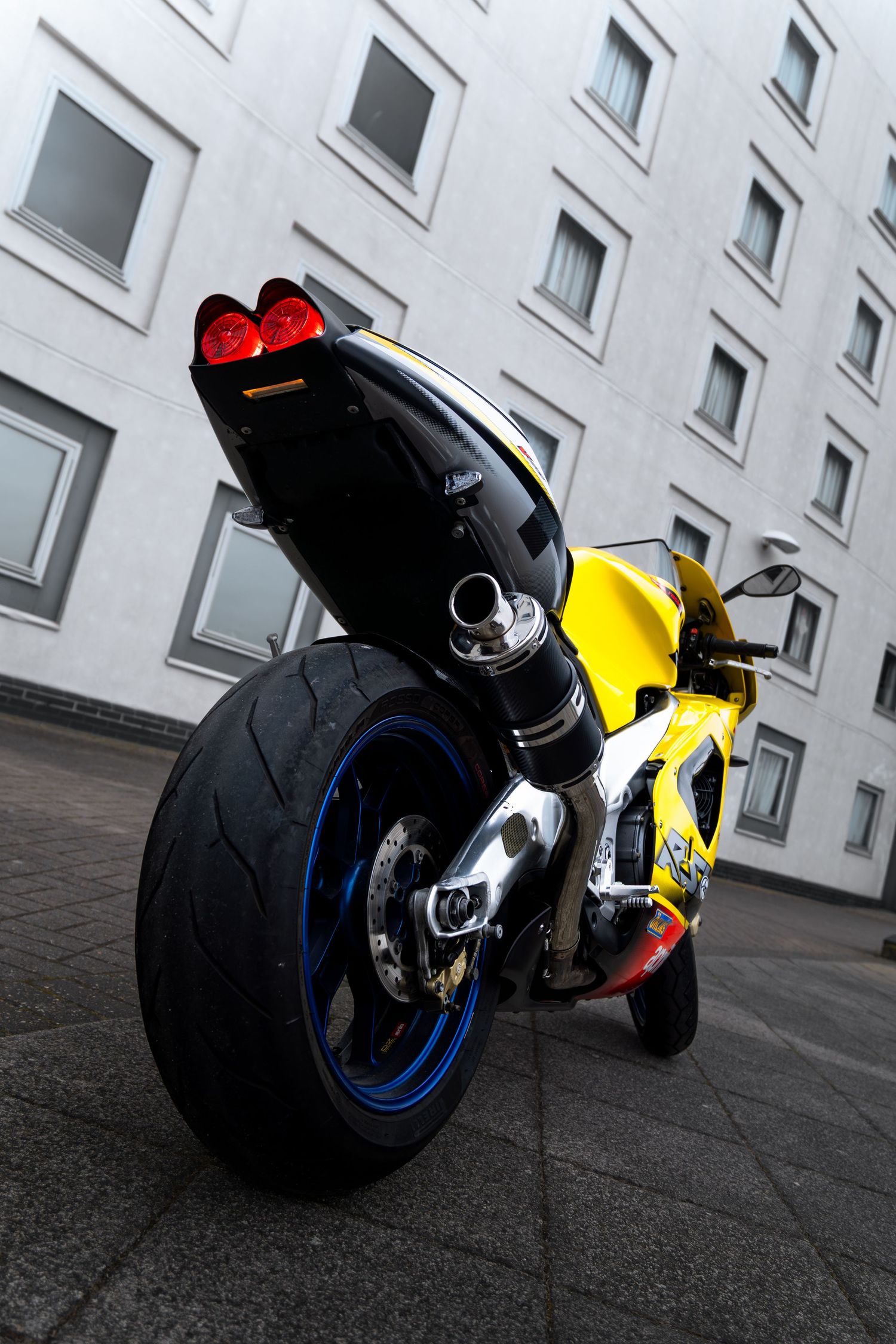 Rear view of yellow Aprilia sport motorcycle with blue wheels and red tail lights illuminated.