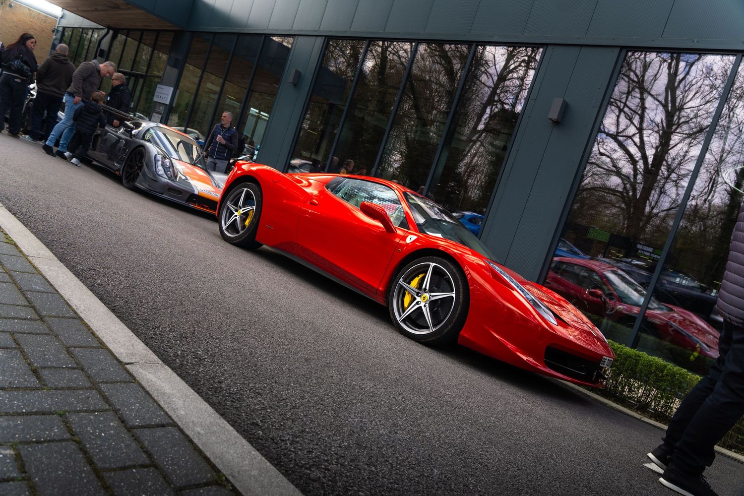 Red Ferrari 458 Italia parked beside Pagani supercar outside glass-fronted luxury car dealership.