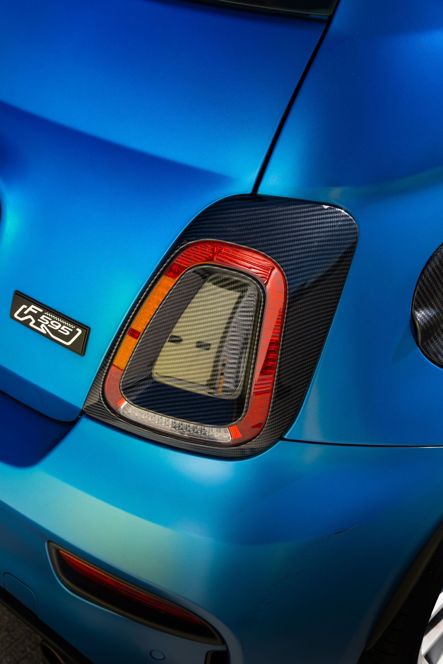 Rear tail light of a blue Abarth 595 with carbon fiber surround and model badge on the bodywork.