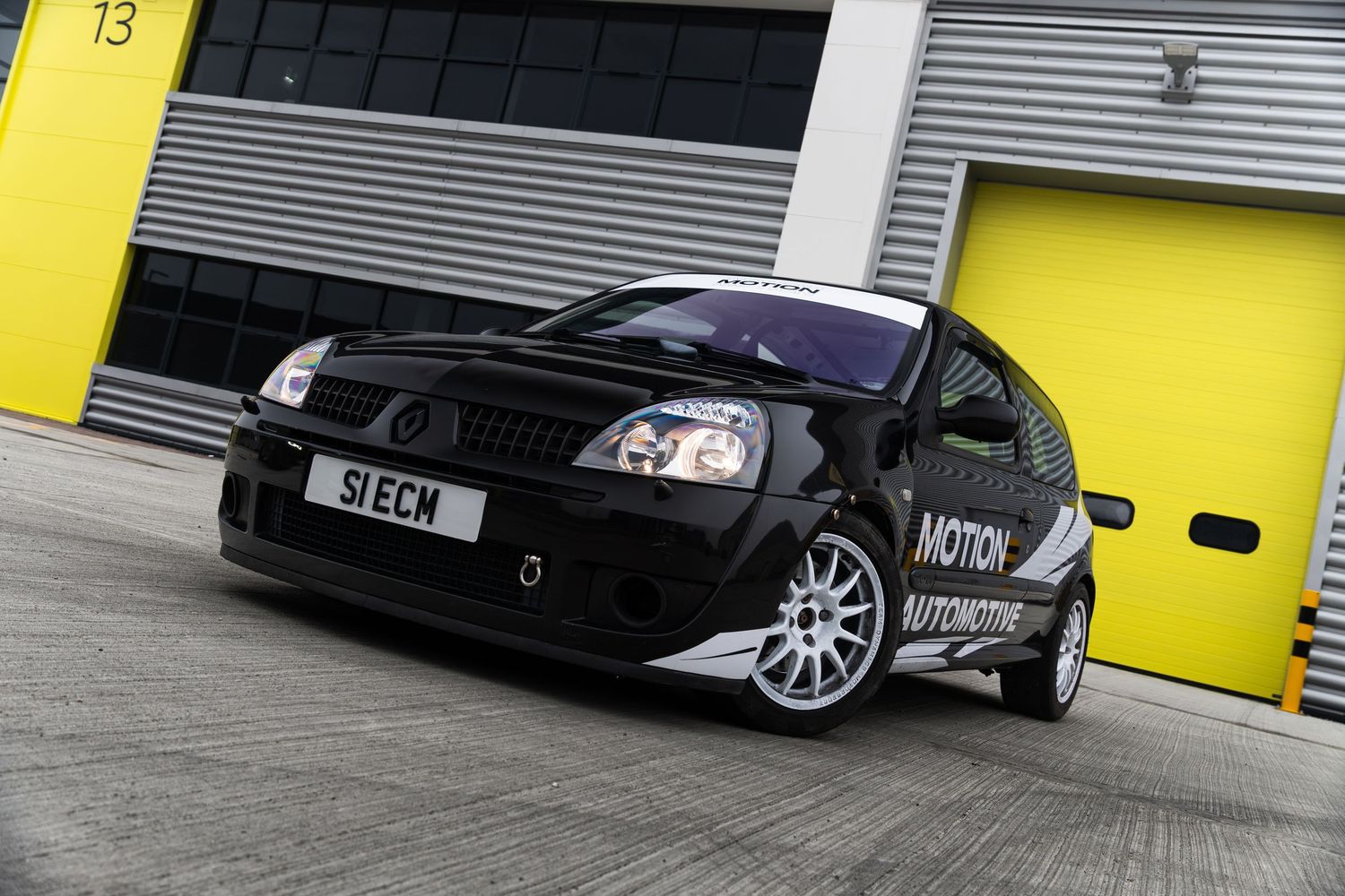 Black Motion Automotive Renault Clio race car with S1ECM number plate parked outside yellow roller shutter doors.