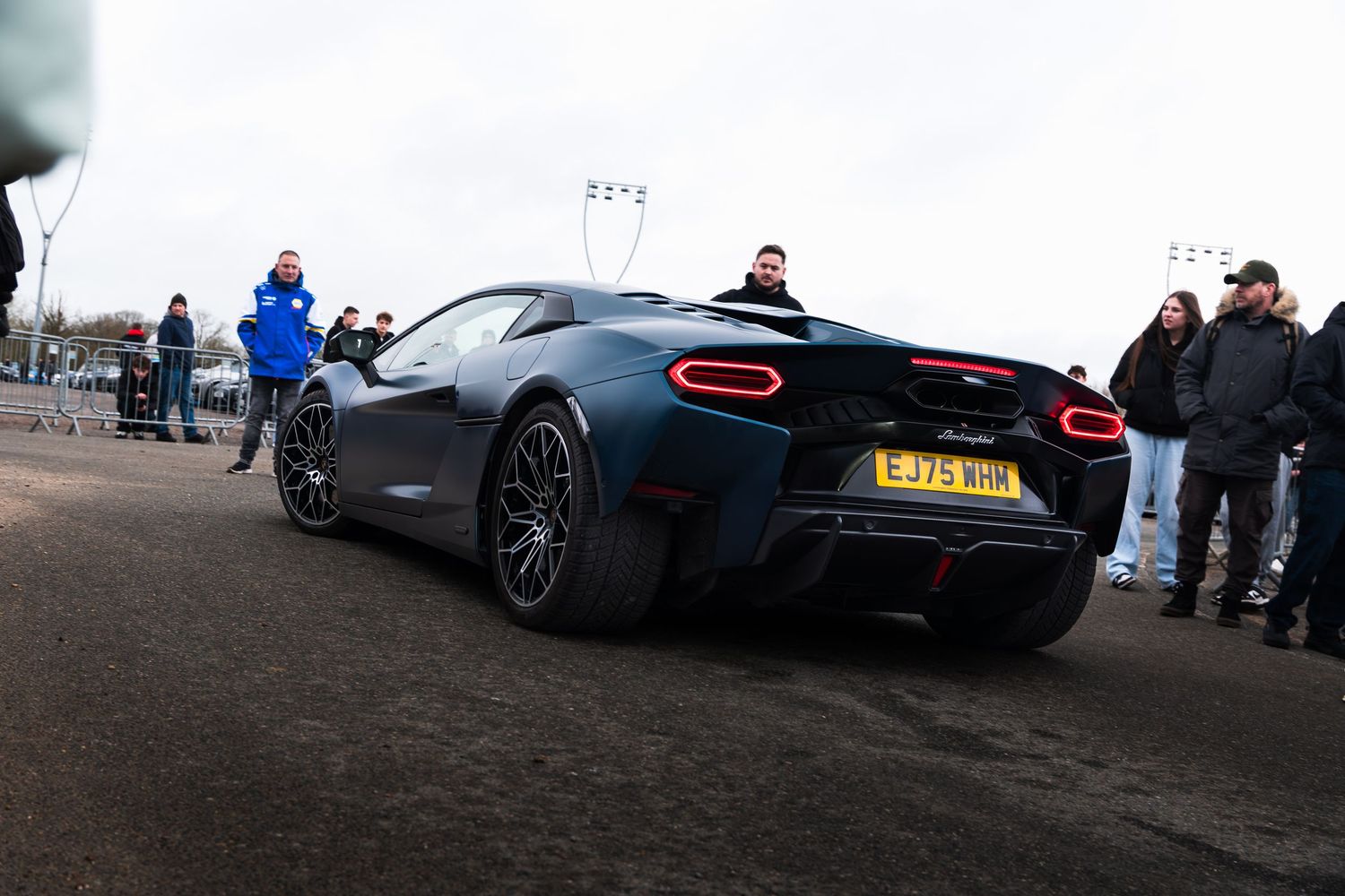 A dark grey McLaren supercar viewed from the rear surrounded by spectators at a car meet.