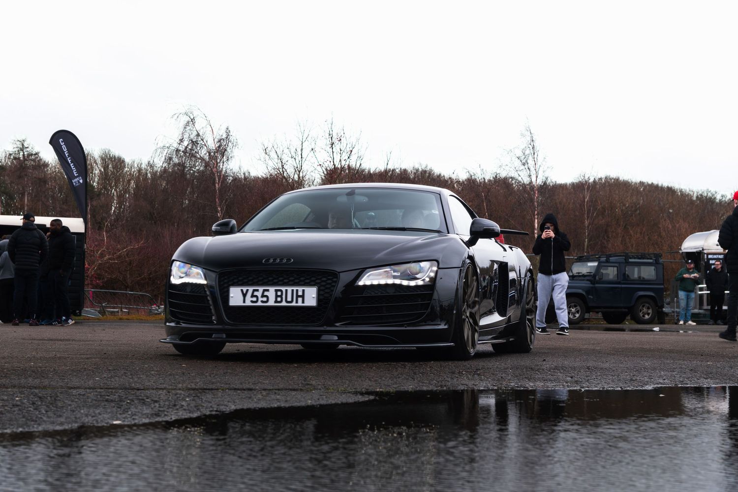 Matte black Audi R8 sports car parked on wet tarmac at an outdoor automotive event.