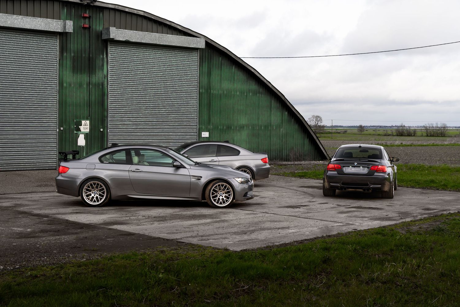 Two BMW M3 E92 coupes parked on concrete in front of a large curved green metal hangar.