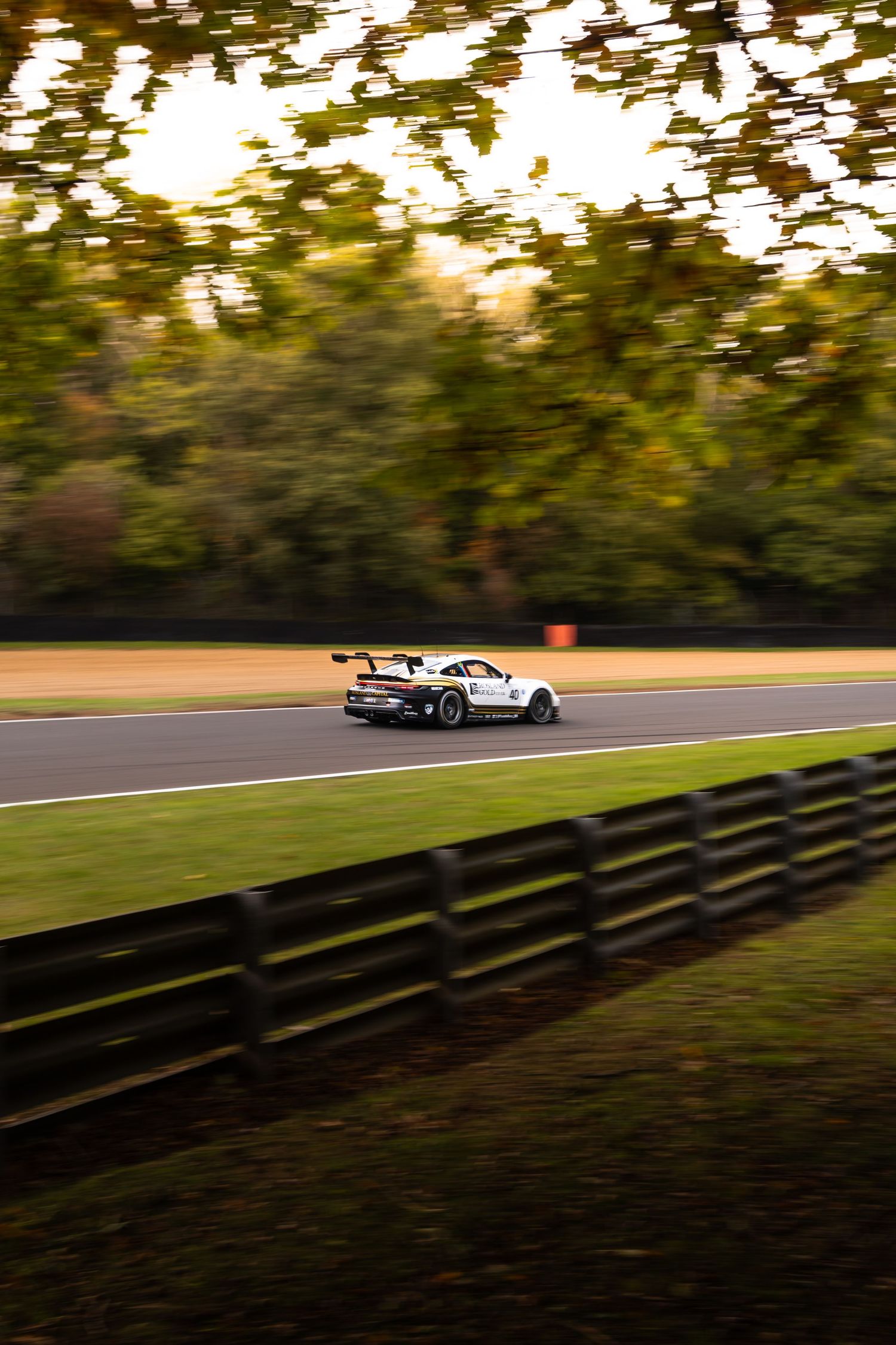 White Porsche GT3 race car speeding on track with autumn trees and wooden fence in background.