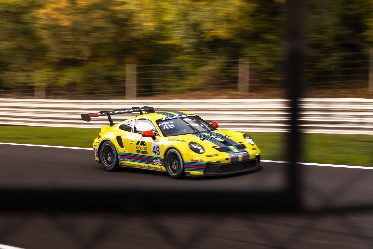 Yellow Porsche 911 GT3 race car with green stripe speeding past, shot through a fence with motion blur.