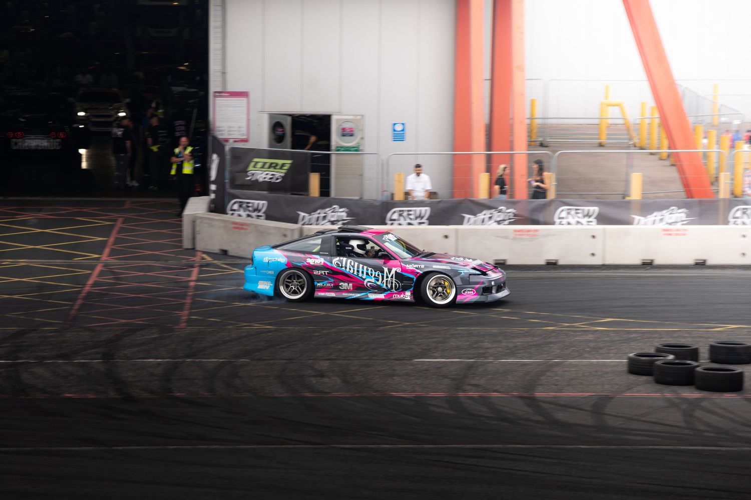 A pink and black drift car slides through a turn generating tire smoke at an indoor motorsport arena event.