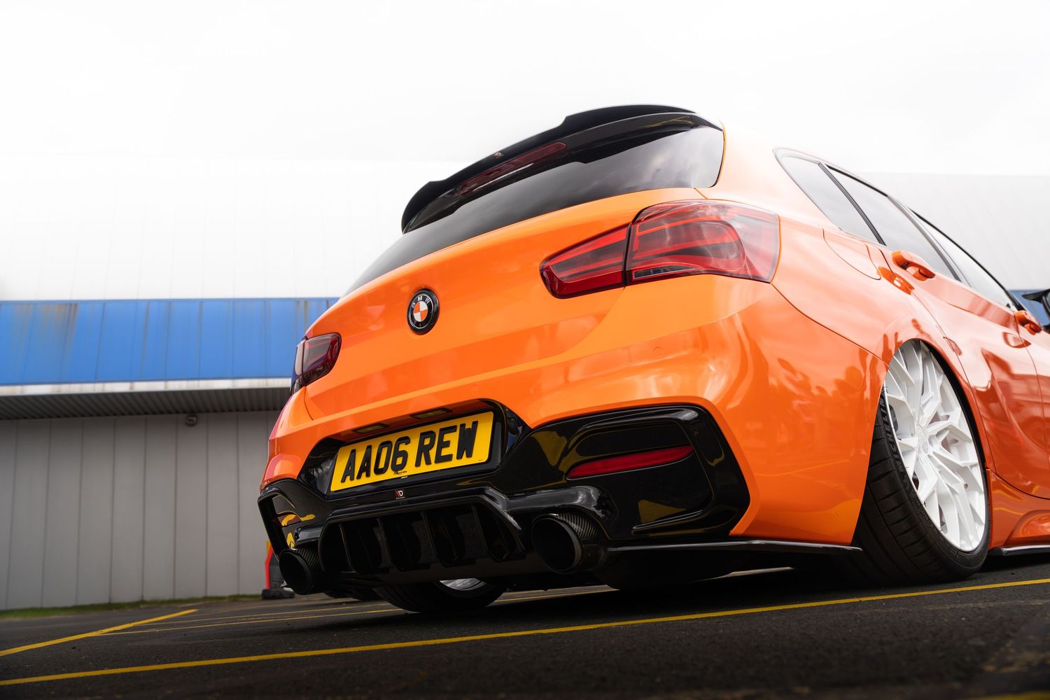Low angle rear view of a modified orange BMW with diffuser and AA06 REW plate at a race track.