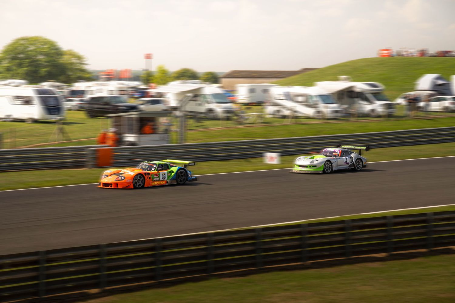 Two GT racing cars competing on a circuit with camper vans in background.
