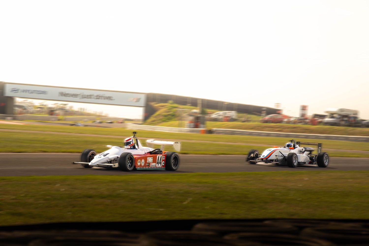 Two Formula racing cars battle side by side on a sunlit motorsport circuit during a race.